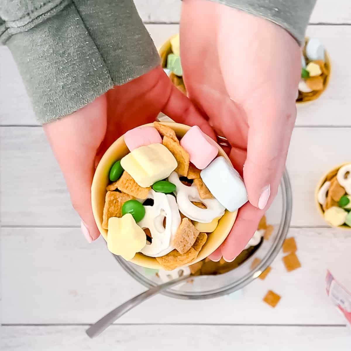A person holds a small yellow bowl filled with cereal squares, green candies, pretzels, and pastel marshmallows over a table.