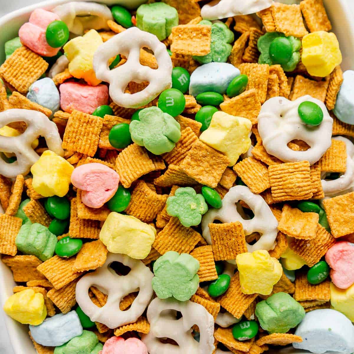 A close-up of a snack mix containing frosted pretzels, green candy-coated chocolates, colorful marshmallows, and square golden cereal pieces.