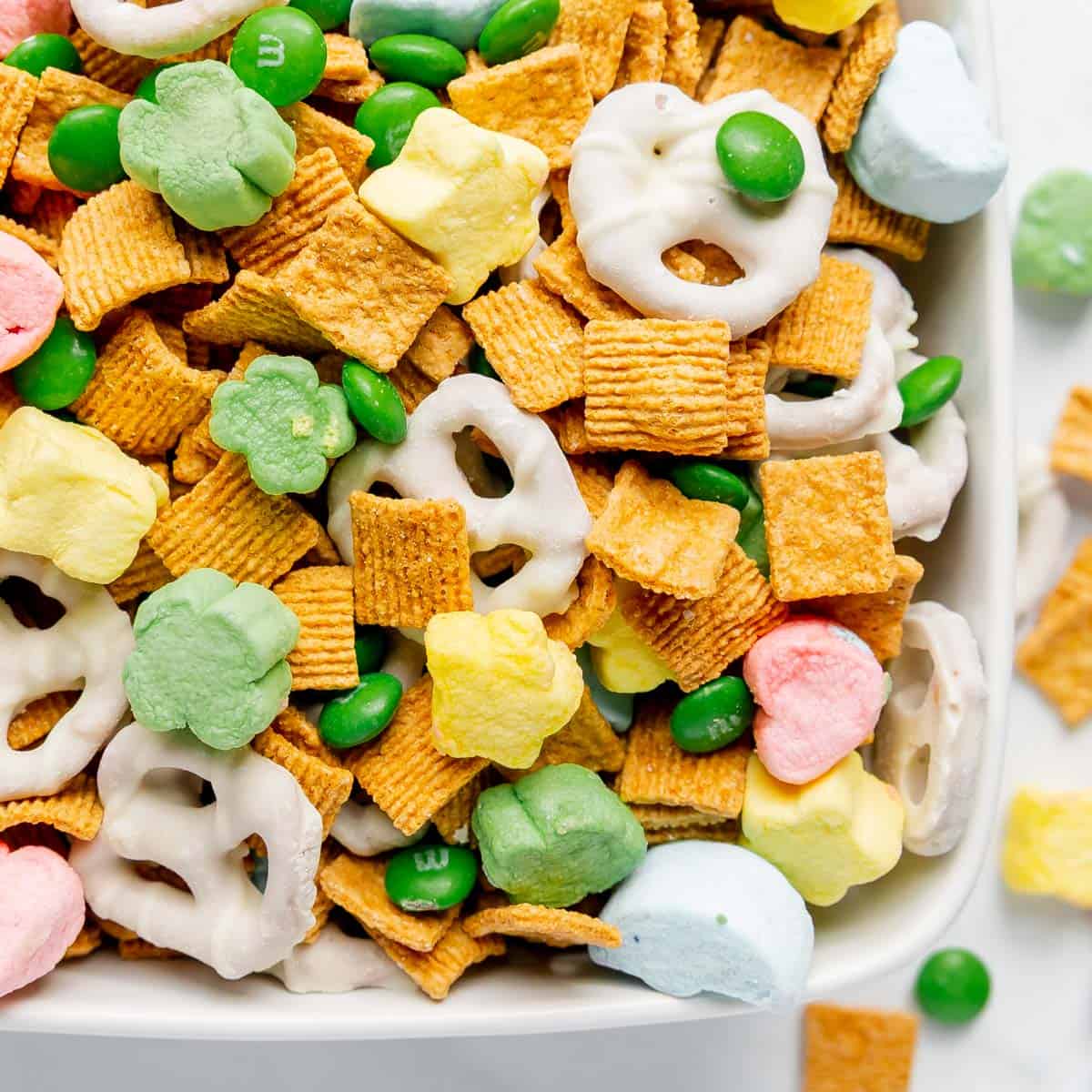 A bowl filled with frosted pretzels, green candies, colorful marshmallows, and square-shaped cereal pieces.