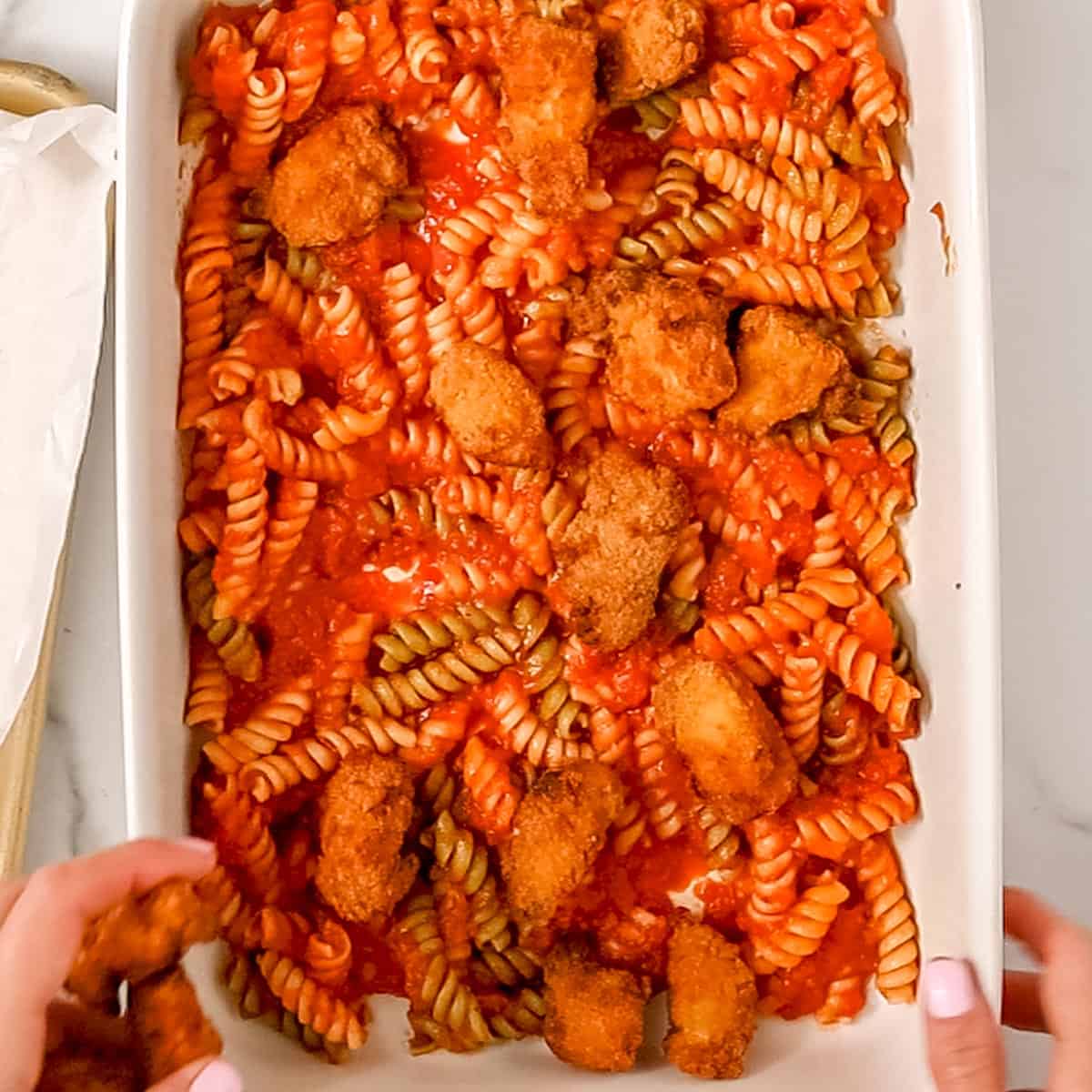 A baking dish filled with tri-color rotini pasta, marinara sauce, and breaded chicken pieces, with a pair of hands adding more chicken.