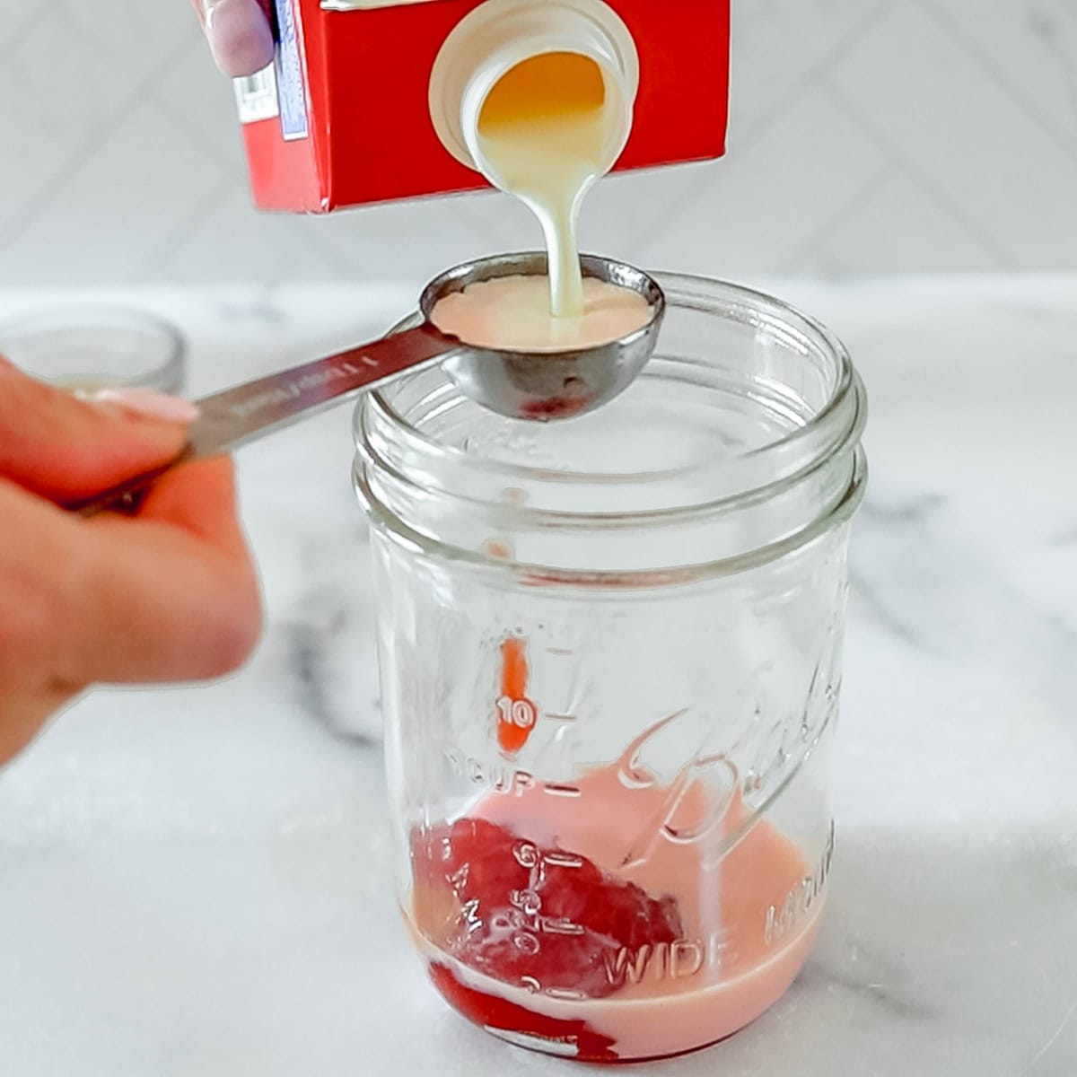 A hand pours cream from a carton into a tablespoon over an empty glass jar, with some cream already inside the jar.