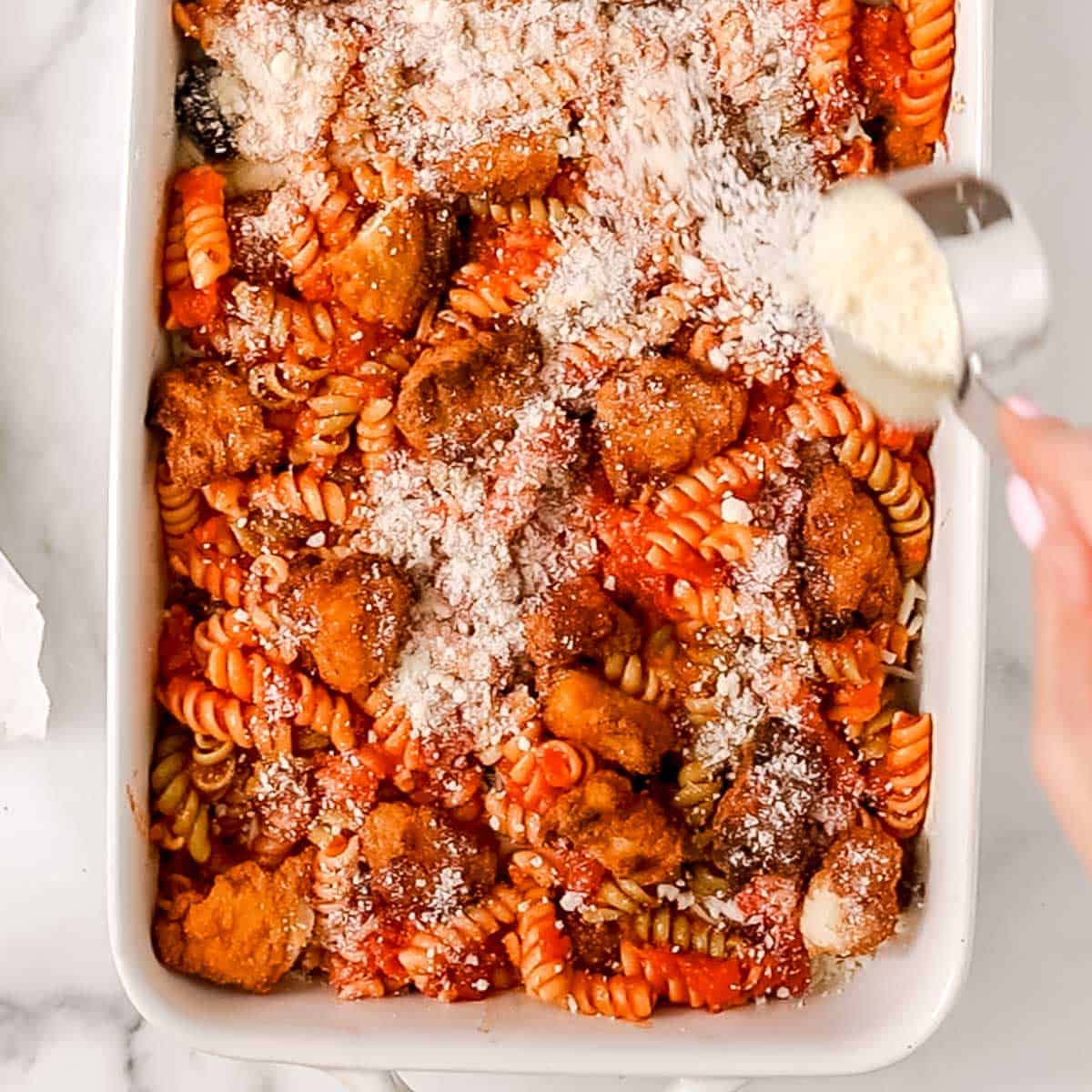 A hand sprinkles grated cheese over a baked pasta dish with rotini, tomato sauce, and breaded chicken pieces in a white rectangular baking dish.