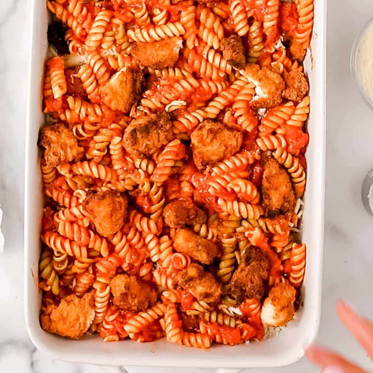 A white baking dish filled with rotini pasta, tomato sauce, and breaded chicken pieces.