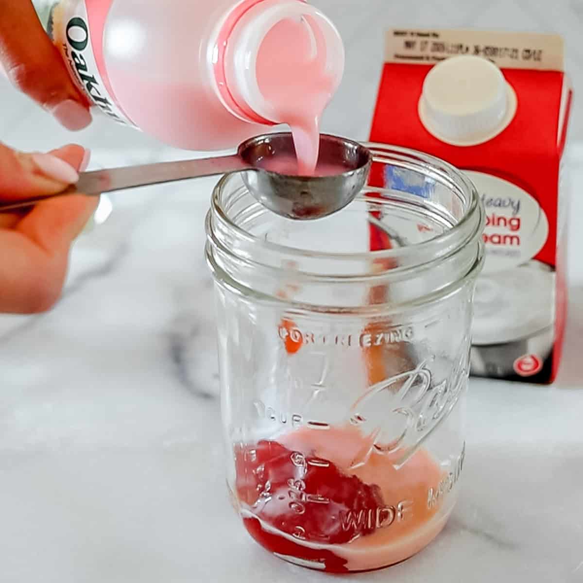 A hand pours pink liquid from a bottle into a measuring spoon over a glass jar, with a carton of heavy whipping cream in the background.