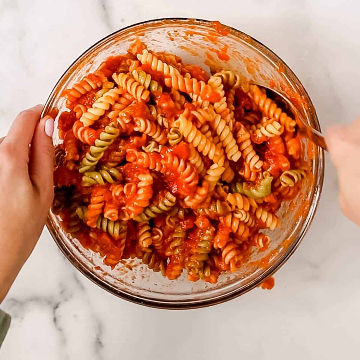 A person mixing tri-color rotini pasta with tomato sauce in a glass bowl on a white marble surface.