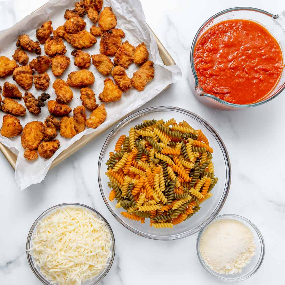 Tray of breaded chicken pieces, a bowl of tri-color rotini pasta, a bowl of shredded cheese, a bowl of grated parmesan, and a bowl of marinara sauce on a white surface.