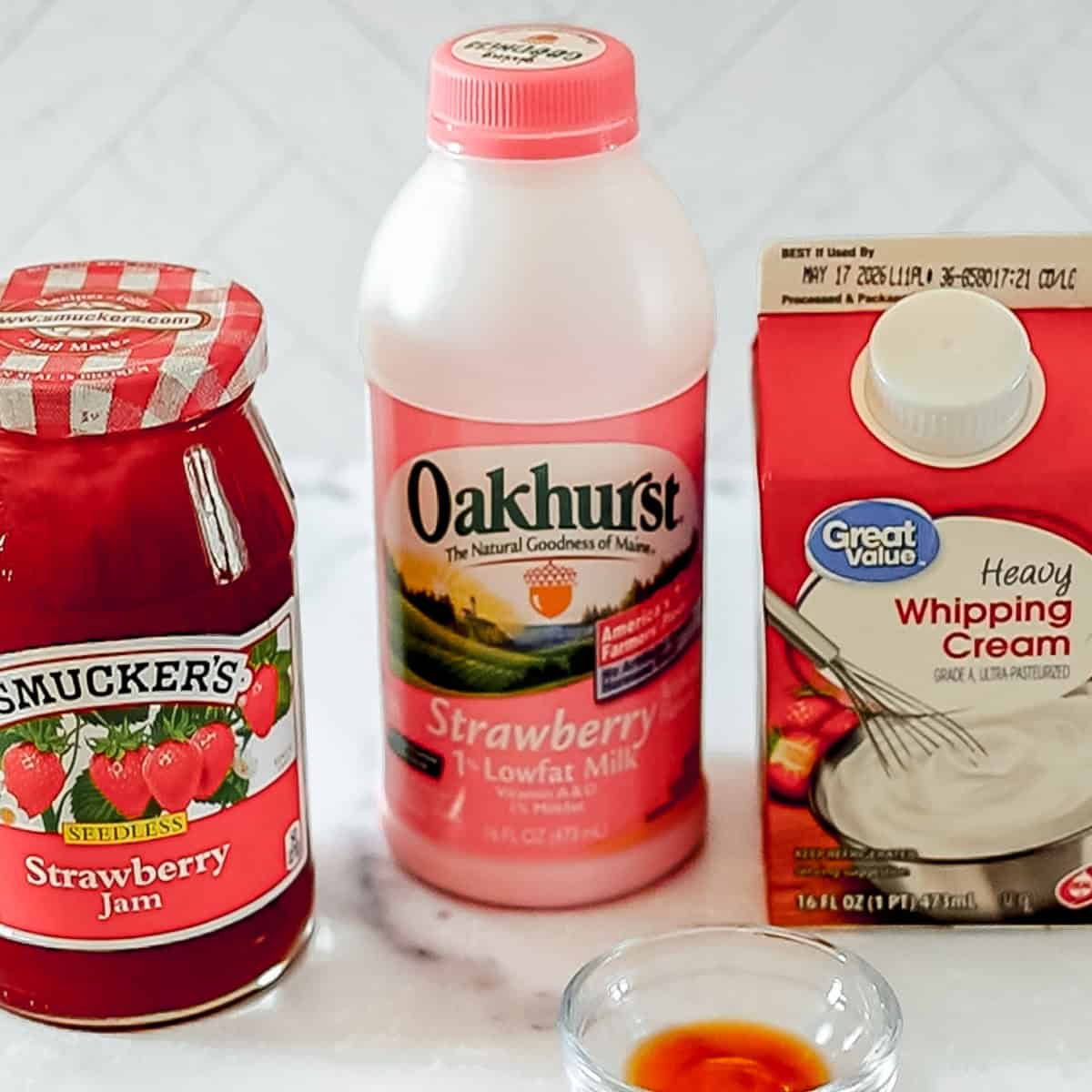 A jar of strawberry jam, a bottle of strawberry low-fat milk, and a carton of heavy whipping cream are placed on a countertop.