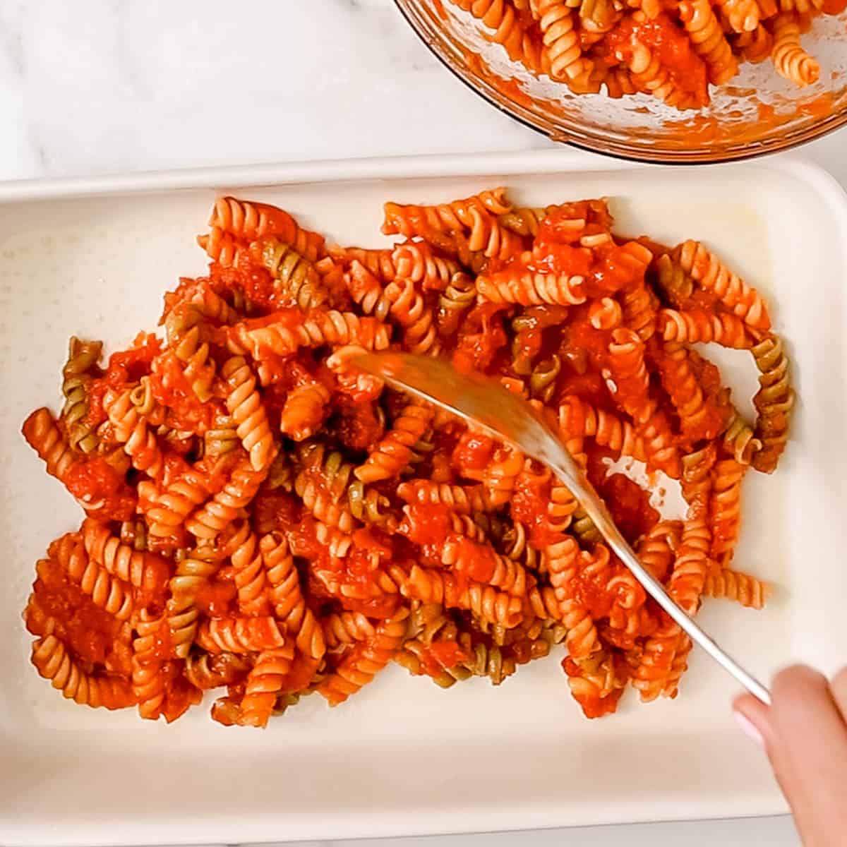 A hand spreads rotini pasta mixed with tomato sauce in a white baking dish, with more pasta visible in a glass bowl nearby.