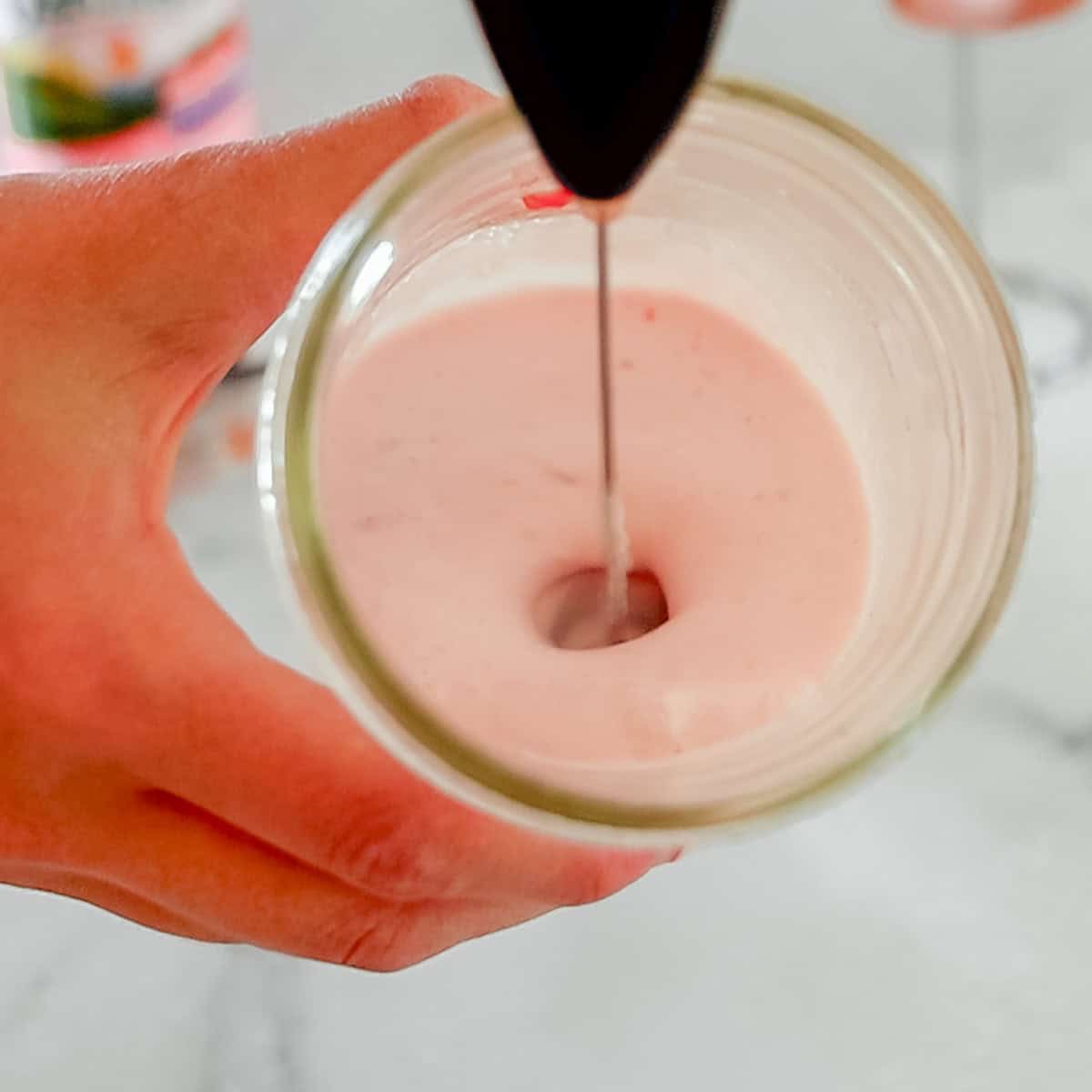 A hand holds a glass filled with pink liquid while a handheld milk frother mixes the contents.