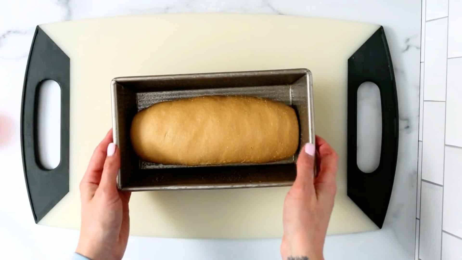 A person’s hands hold a loaf pan with unbaked bread dough on a large cutting board.