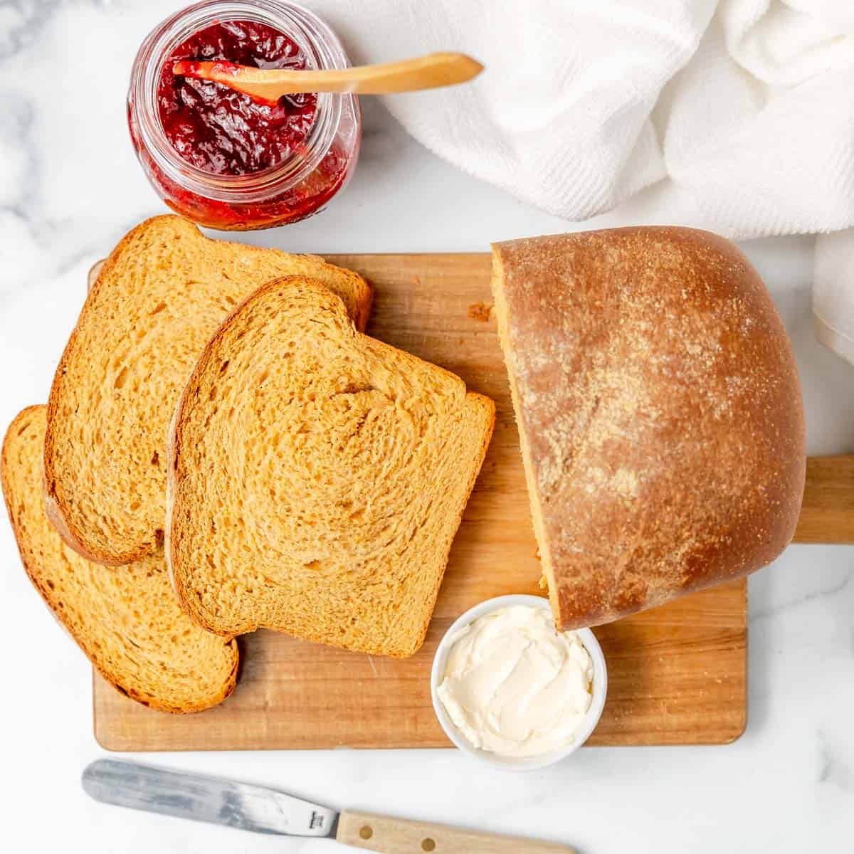 A loaf of bread with three slices, a jar of red jam, a small dish of spread, and a knife on a wooden cutting board.