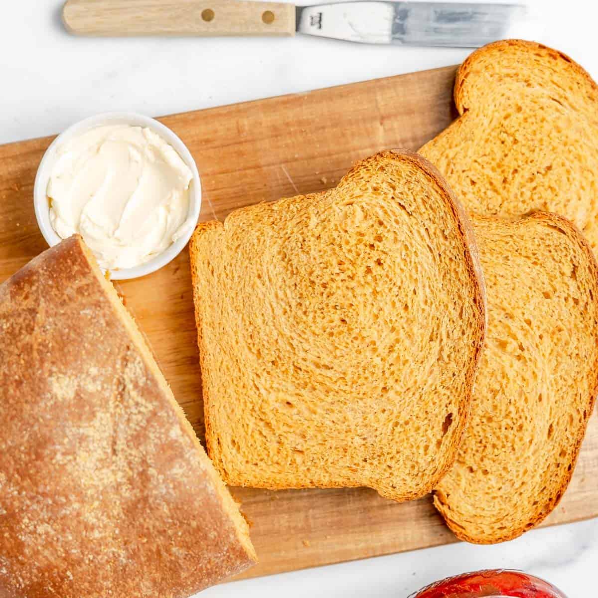 Three slices of bread, a partial loaf, a small dish of butter, and a butter knife are arranged on a wooden cutting board.