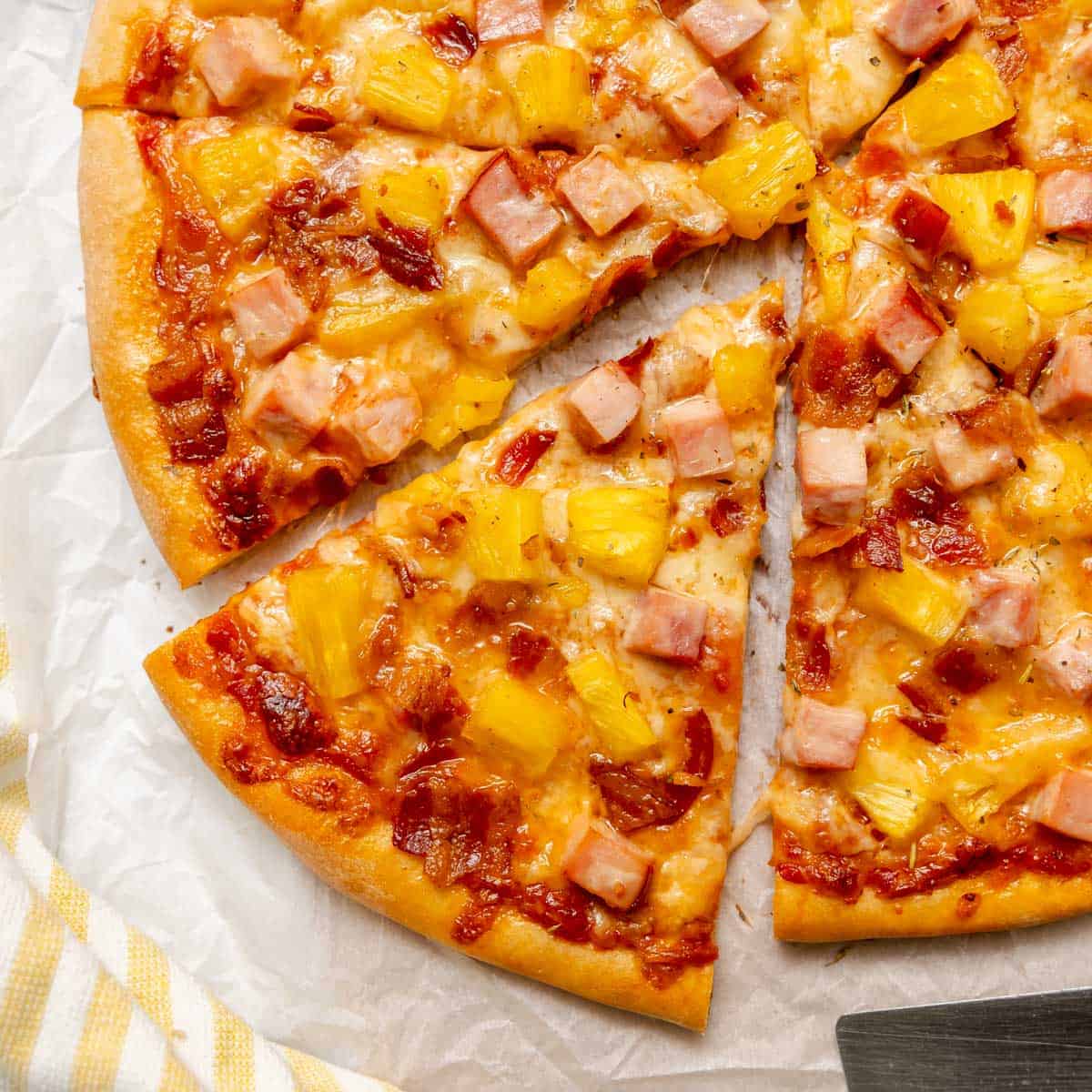 A Hawaiian pizza topped with ham, pineapple, bacon, and melted cheese is sliced and served on parchment paper.