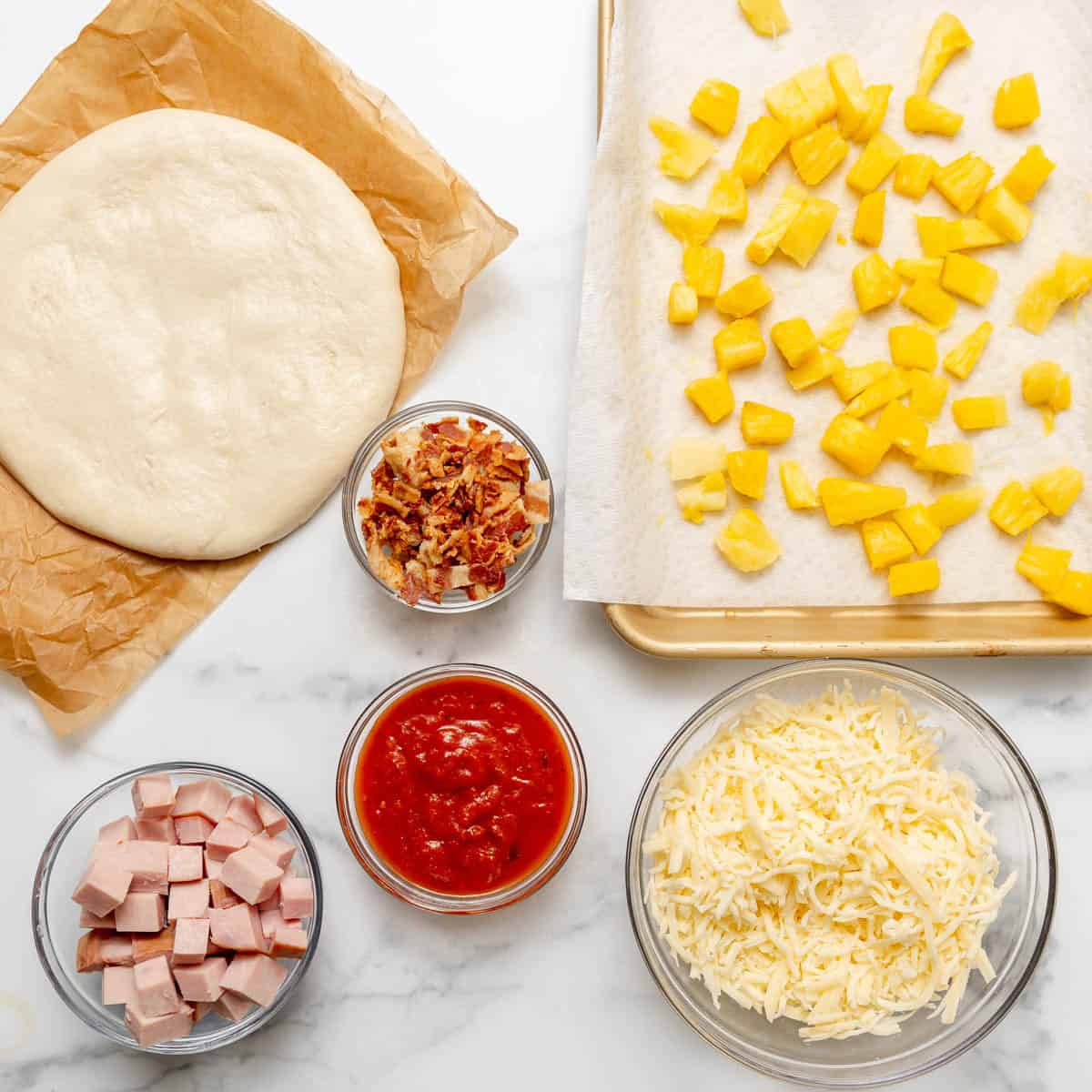 Pizza ingredients on a white surface: dough, chopped pineapple, shredded cheese, red sauce, cubed ham, and cooked bacon bits in separate bowls and containers.