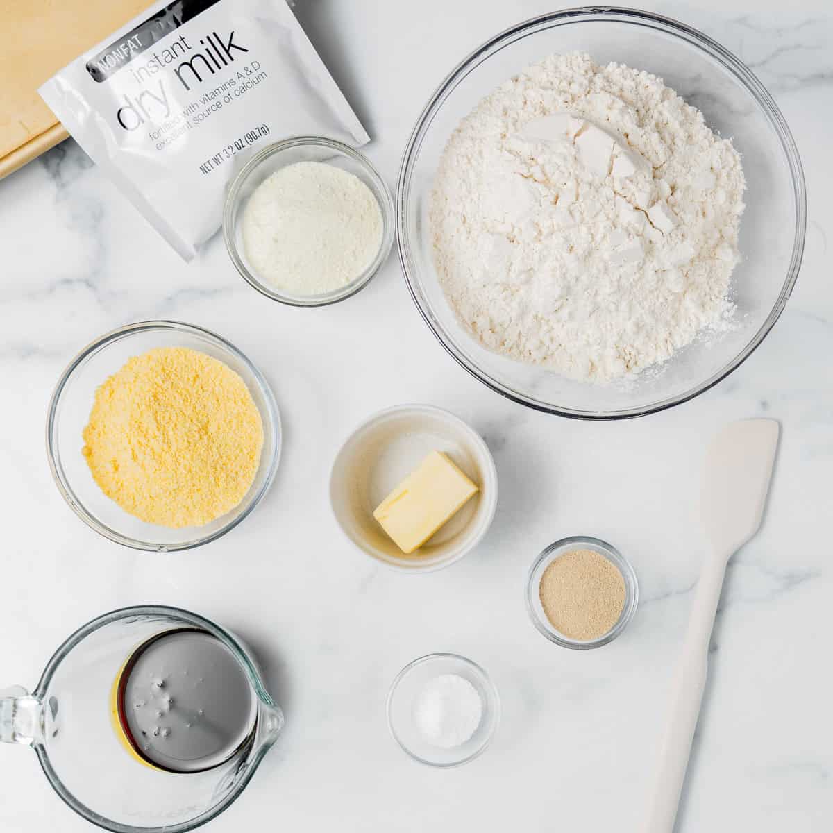Top-down view of baking ingredients on a white surface, including flour, cornmeal, butter, instant dry milk, salt, yeast, molasses, and a white spatula.