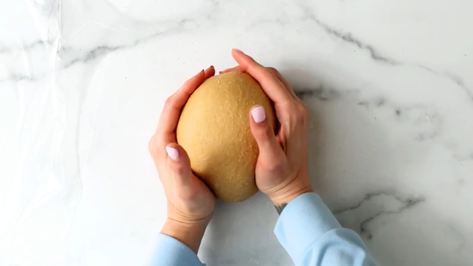 Two hands with light pink nail polish are holding a round ball of dough on a white marble surface.