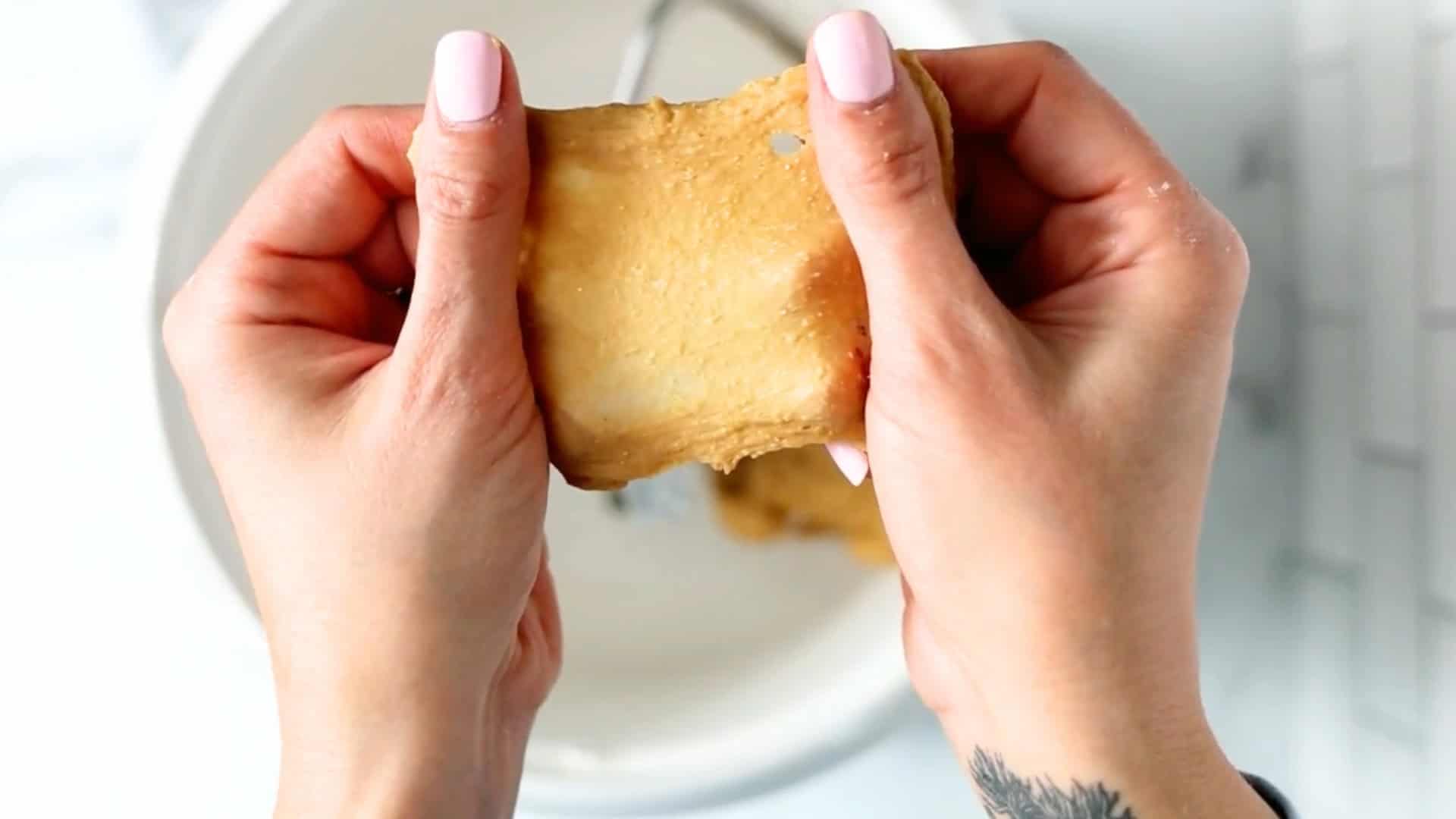 Two hands stretch a piece of tan dough or seitan over a white bowl, showing its texture.