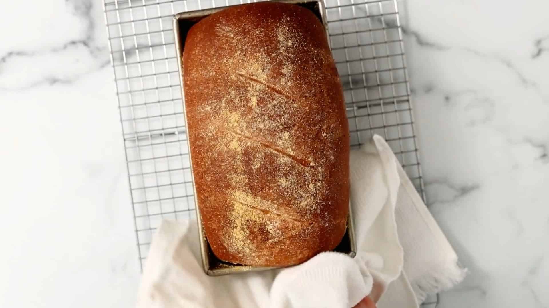 A hand holding a cloth lifts a loaf of golden brown bread in a rectangular pan, set on a wire cooling rack over a marble surface.