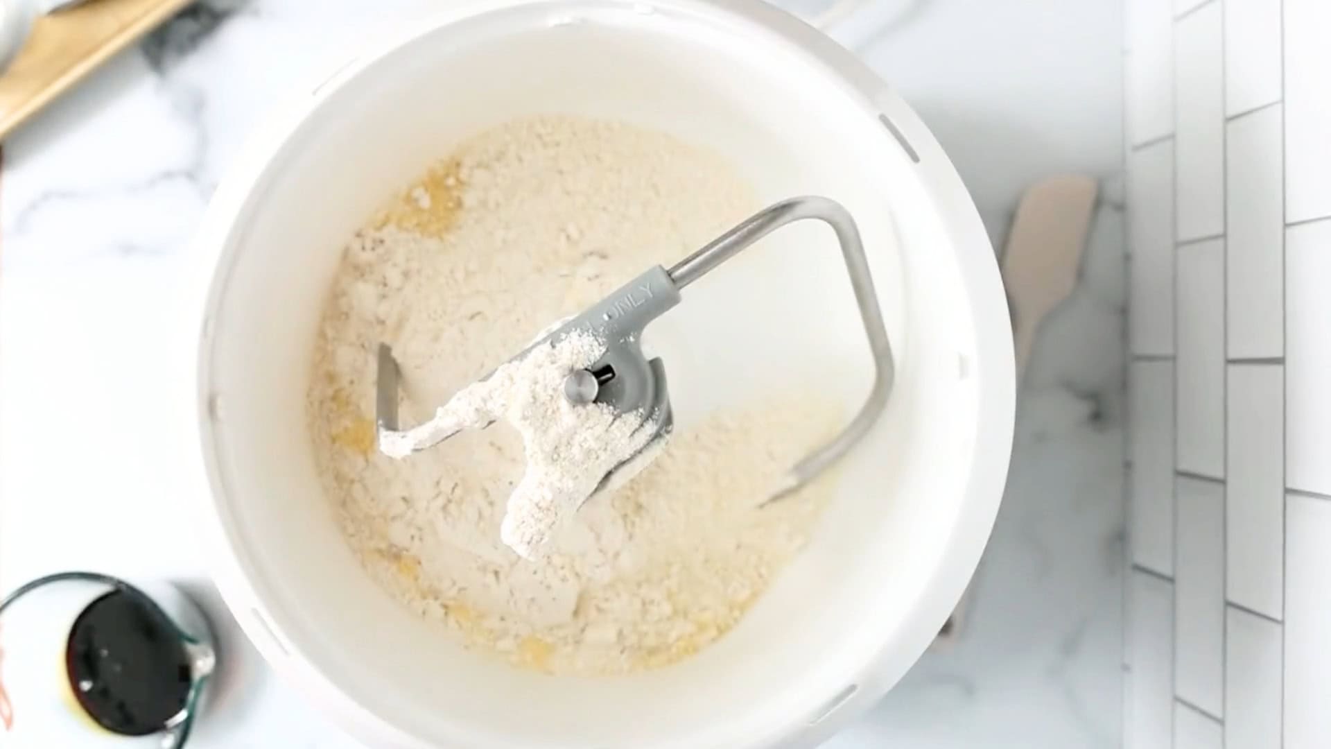 A white mixing bowl containing flour and wet ingredients with a dough hook attachment, ready for mixing homemade Anadama Bread.
