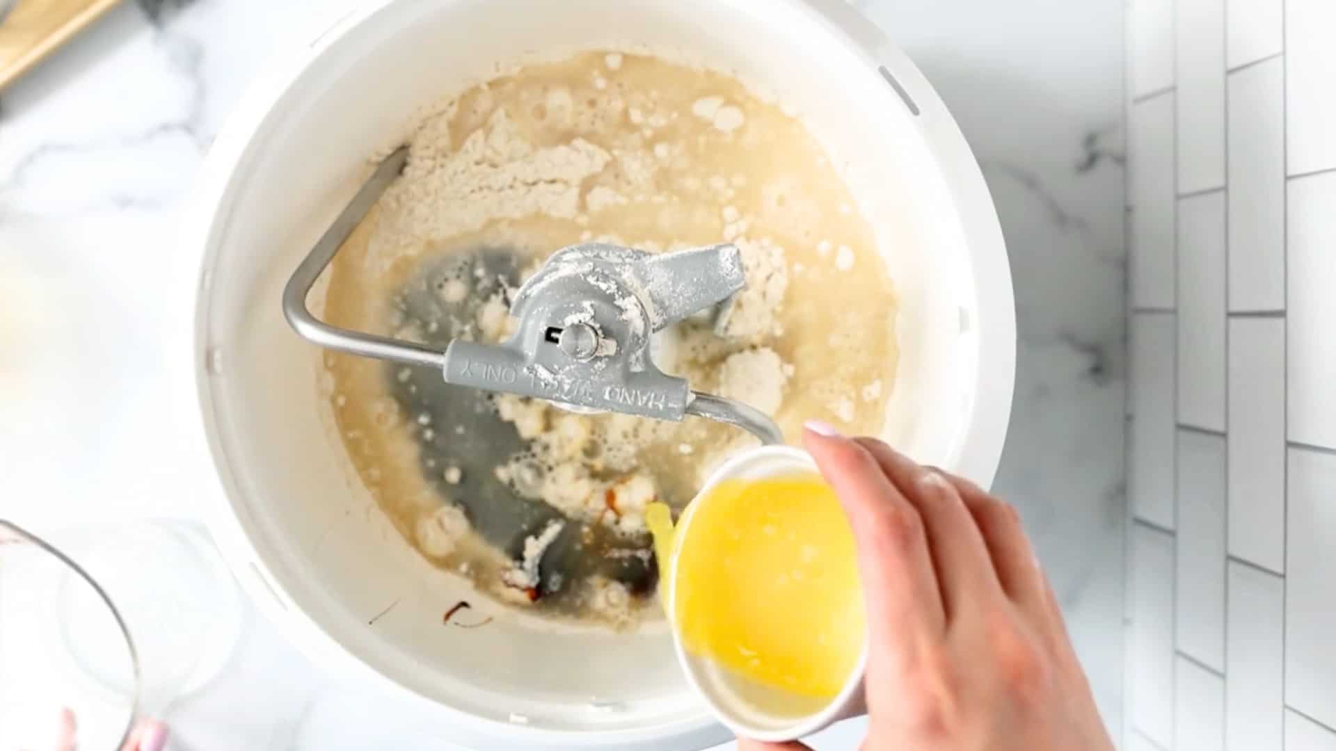 A hand pours melted butter into a stand mixer bowl containing flour, yeast, and other wet ingredients, preparing dough.