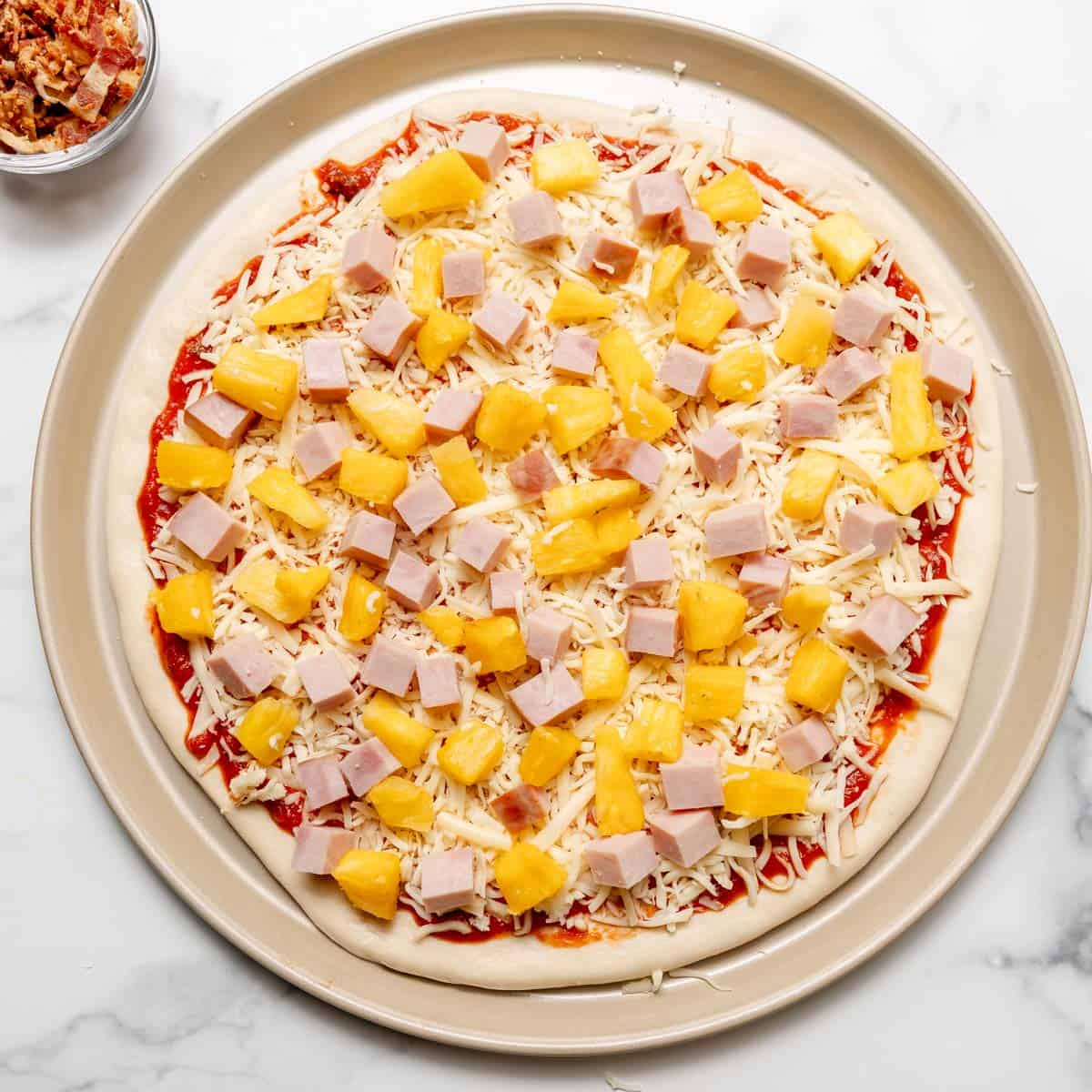 Unbaked pizza topped with tomato sauce, shredded cheese, diced ham, and pineapple chunks on a round baking tray.