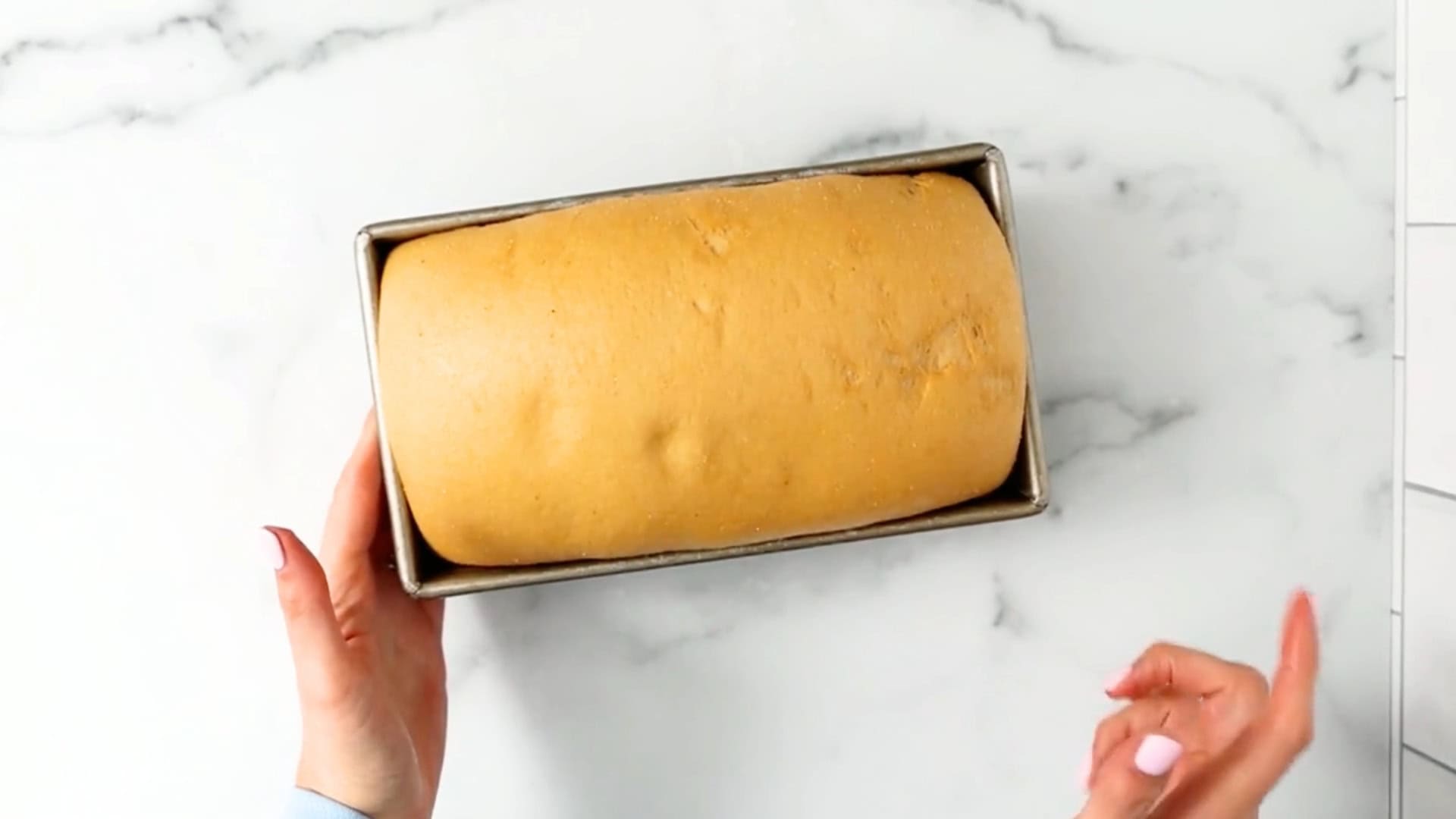 A person holds a loaf of unbaked bread dough in a rectangular metal pan over a white marble surface.