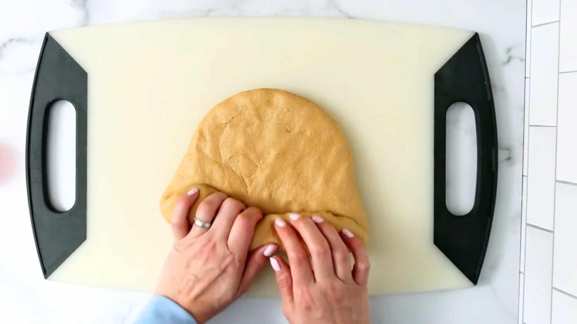 Hands folding a ball of dough on a light-colored cutting board with black handles, on a white countertop.