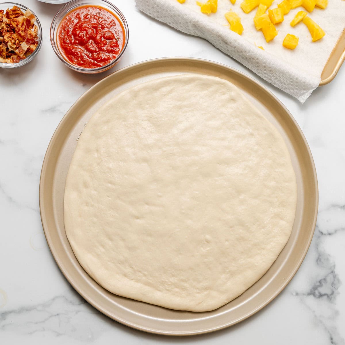 Round pizza dough on a baking pan with bowls of tomato sauce and bacon, and chopped pineapple pieces on a paper towel nearby.