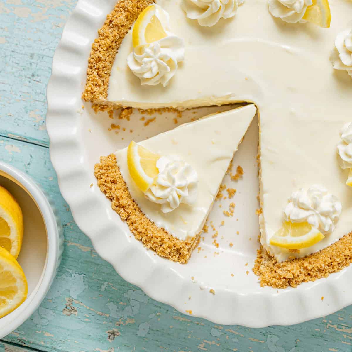 A lemon cream pie with a graham cracker crust, topped with whipped cream and lemon slices, sits on a white plate with one slice removed. A bowl of lemon wedges is beside it.