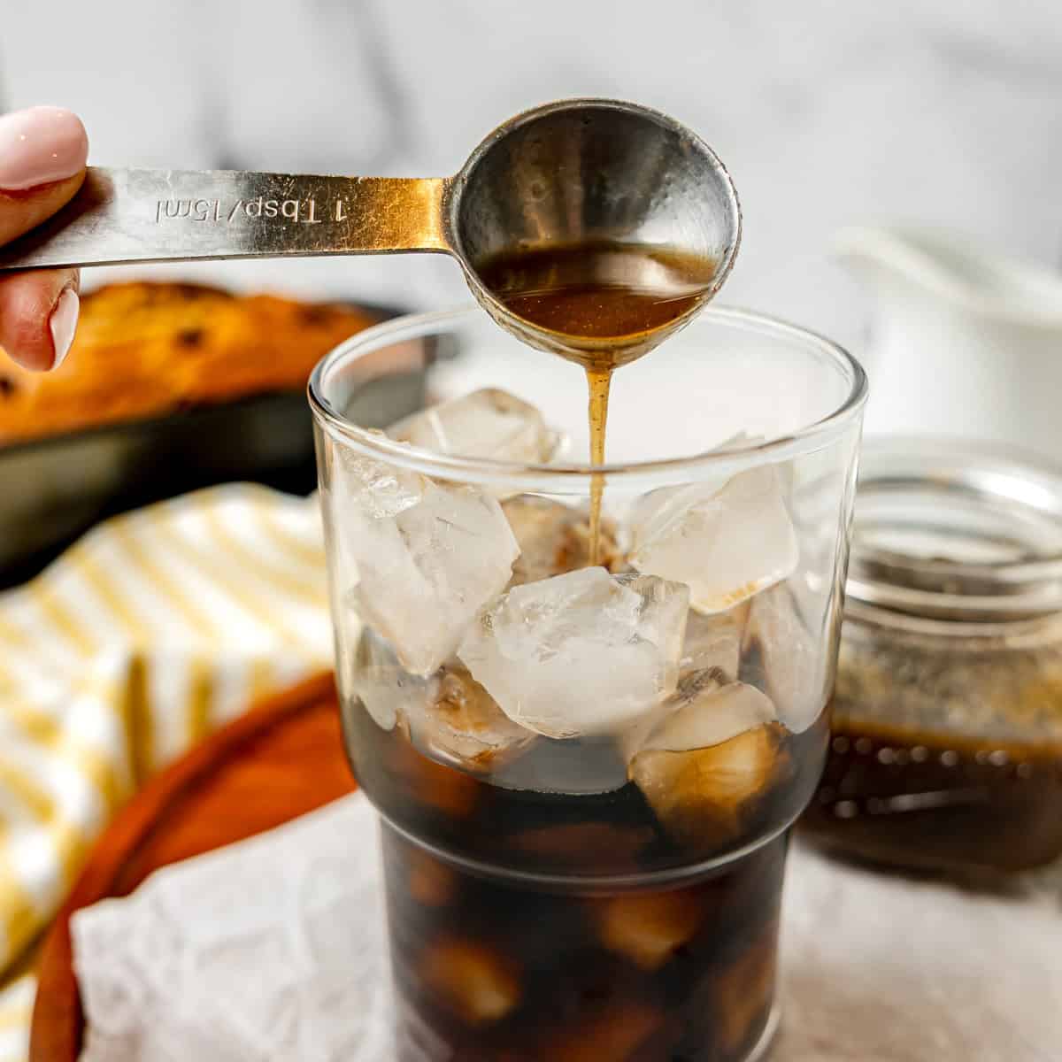 A hand pours liquid from a measuring spoon into a glass filled with ice and dark beverage; a jar and cloth are in the background.