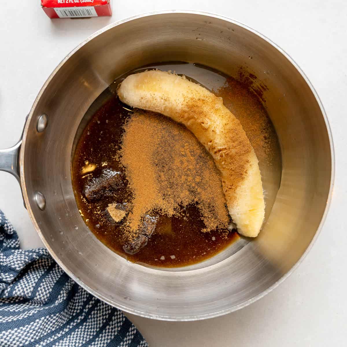 A saucepan containing a whole banana, brown sugar, cinnamon, and liquid ingredients, placed on a white surface next to a blue-striped towel.
