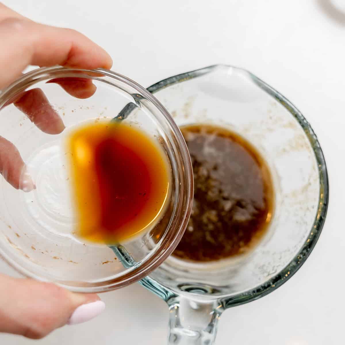 A hand holds a small glass bowl with brown liquid over a larger measuring cup containing a similar brown liquid.