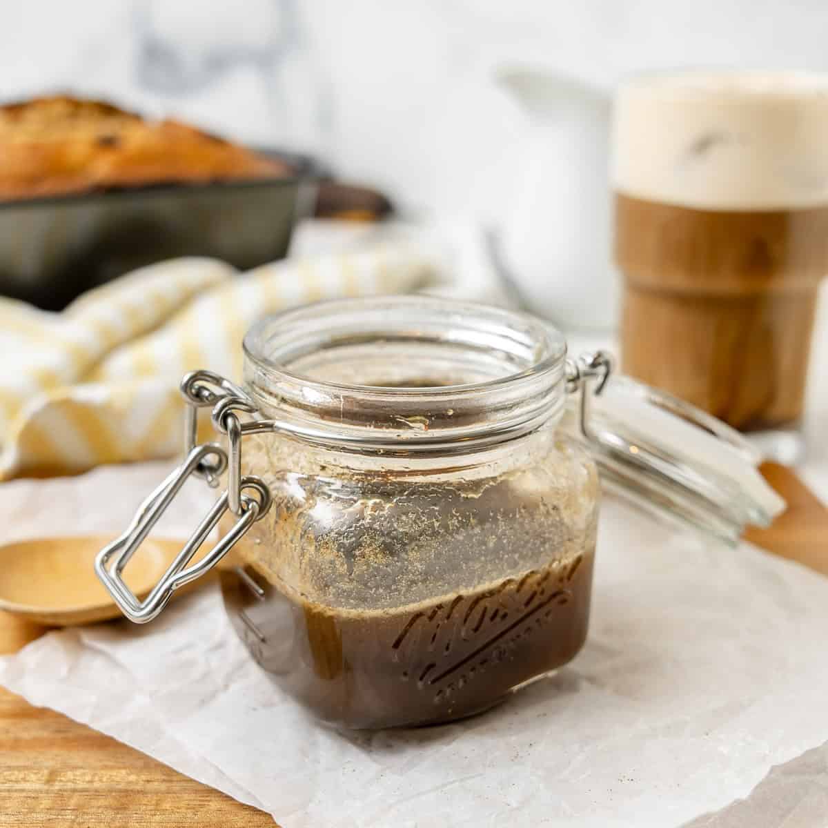 A glass jar filled with brown syrup sits on parchment paper, with a wooden spoon, a loaf of bread, and a glass of iced coffee in the background.
