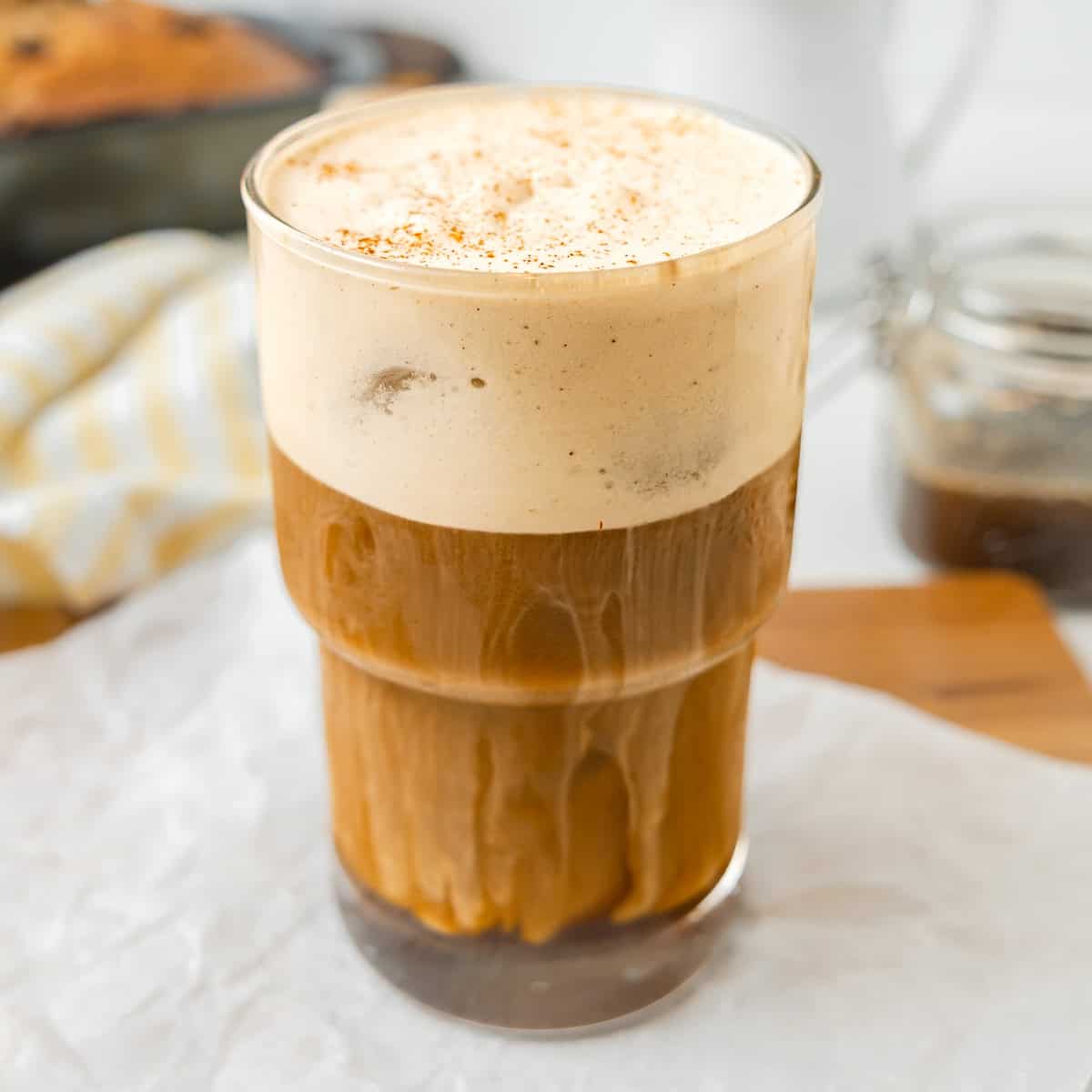A glass of iced coffee with a thick layer of frothy cream on top, sitting on a white surface with a jar and striped cloth in the background.