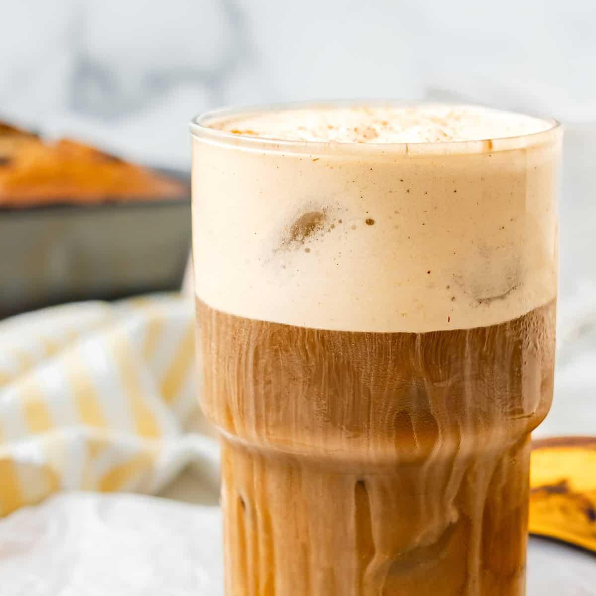 A glass of iced coffee with creamy foam on top, placed on a white surface with a blurred background.