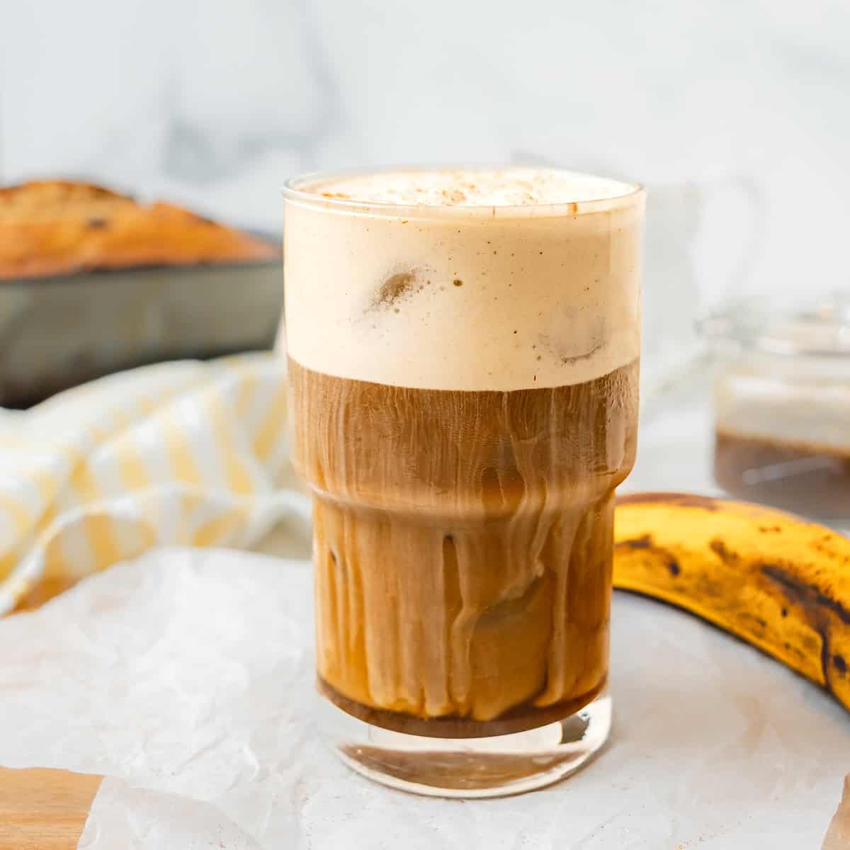 A glass of iced coffee with creamy foam on top sits on a white surface, with a banana and a baked loaf in the background.