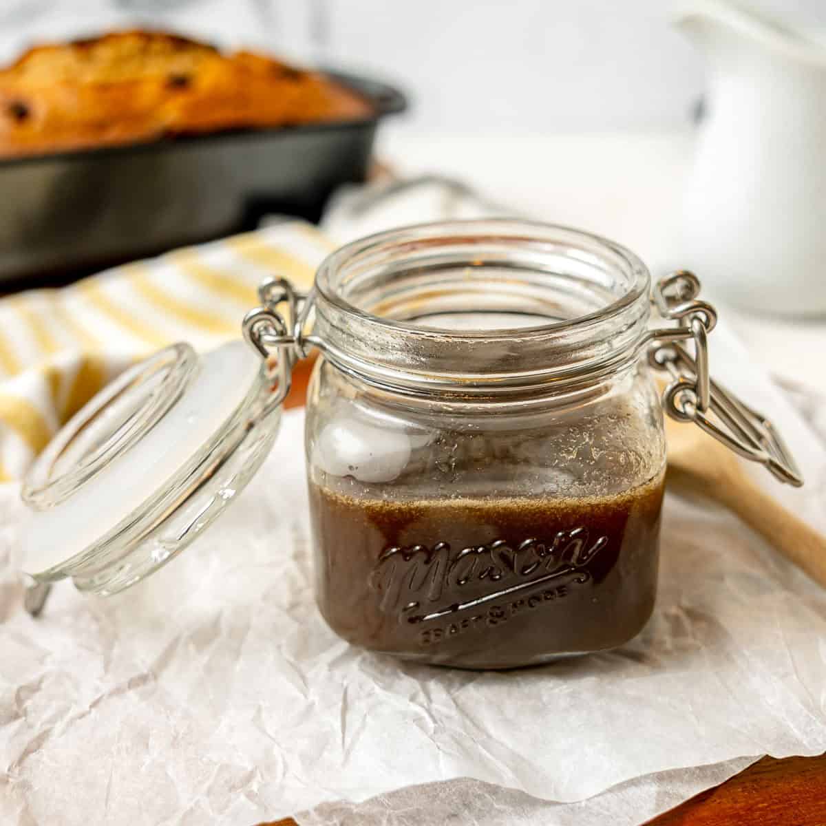 An open glass jar filled with brown syrup sits on parchment paper, with a loaf of bread and a striped cloth in the background.