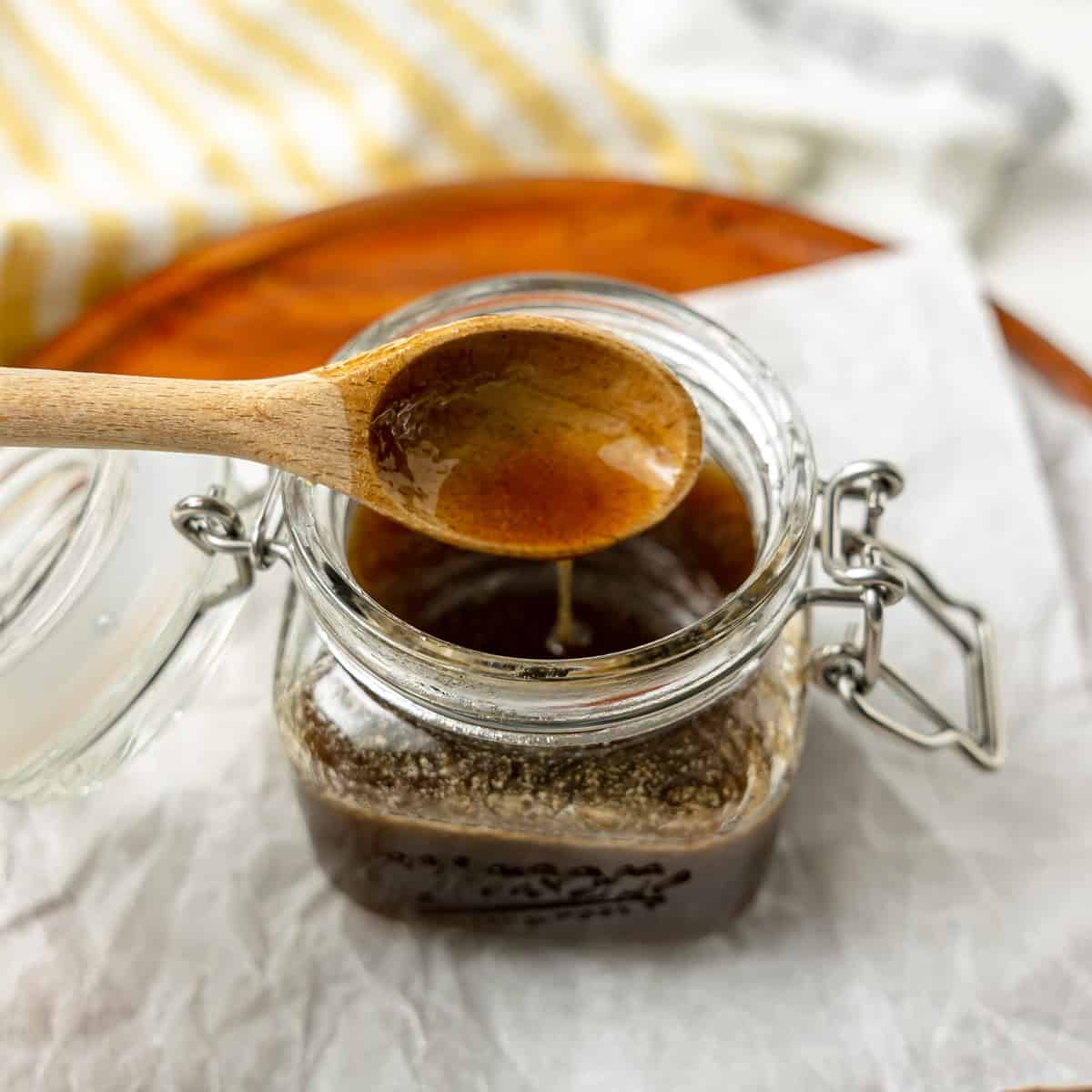 A wooden spoon holds brown liquid above an open glass jar filled with a thick, dark sauce or syrup, placed on parchment paper.