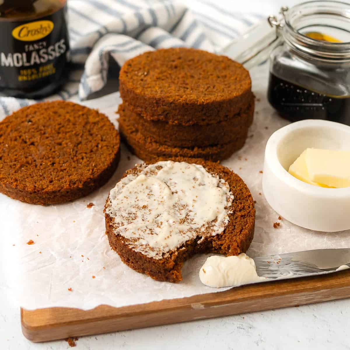 Sliced brown bread with one piece spread with butter, a bite taken out, and jars of molasses and butter on a wooden board.