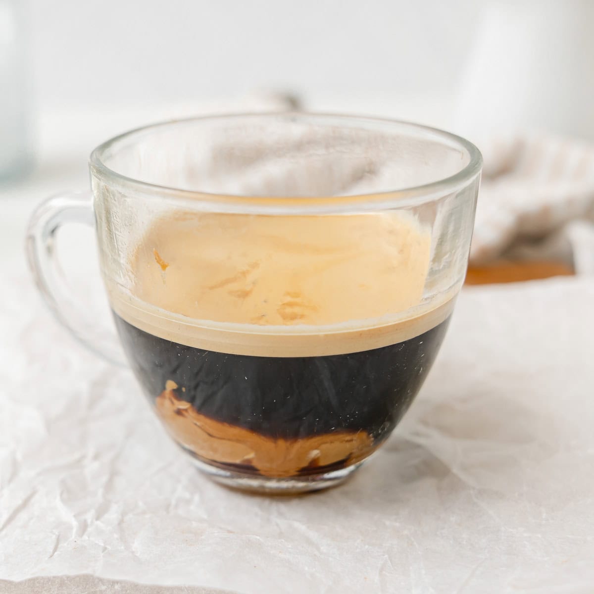 A clear glass cup filled with black coffee and a visible layer of cookie butter at the bottom, placed on a crumpled white surface.