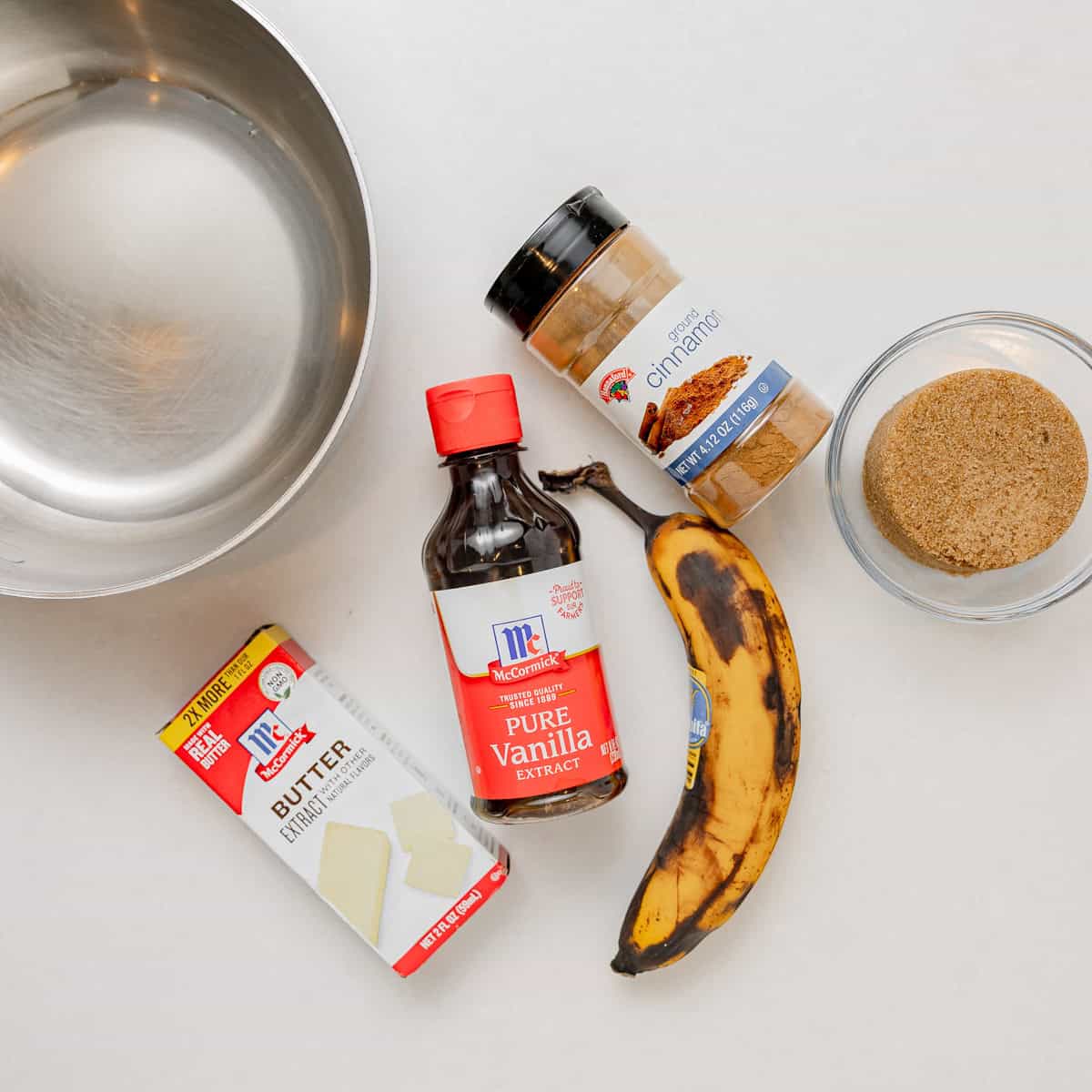 A mixing bowl, a stick of butter, vanilla extract, ground cinnamon, a ripe banana, and a bowl of brown sugar are arranged on a white surface.