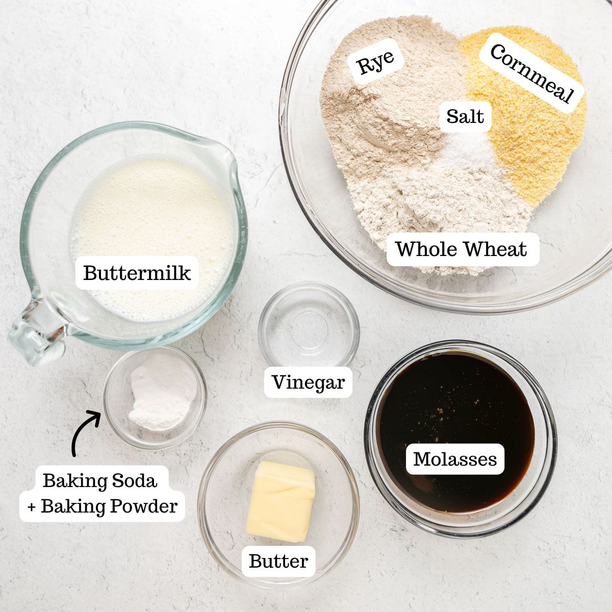 Top-down view of labeled ingredients for baking Boston Brown Bread: rye flour, cornmeal, whole wheat flour, salt, buttermilk, baking soda and powder, vinegar, butter, and molasses on a white surface.