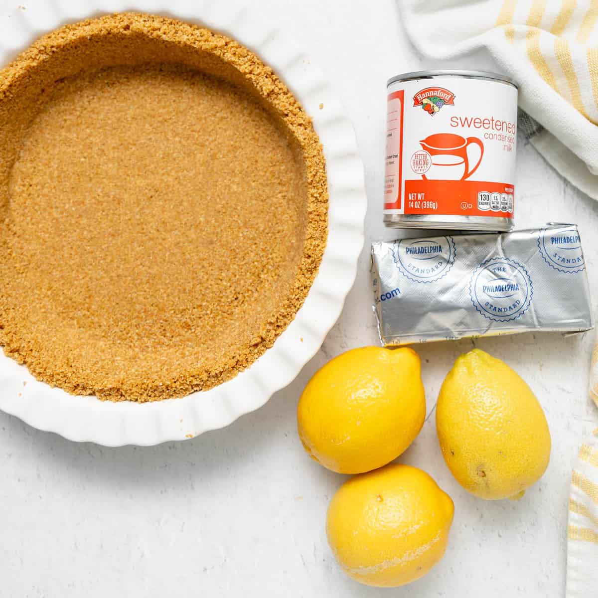 A graham cracker pie crust, a can of sweetened condensed milk, a block of cream cheese, and three lemons on a white surface.