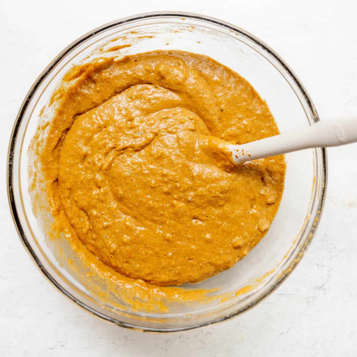 A glass bowl filled with brown bread batter and a white spatula resting inside on a white surface.