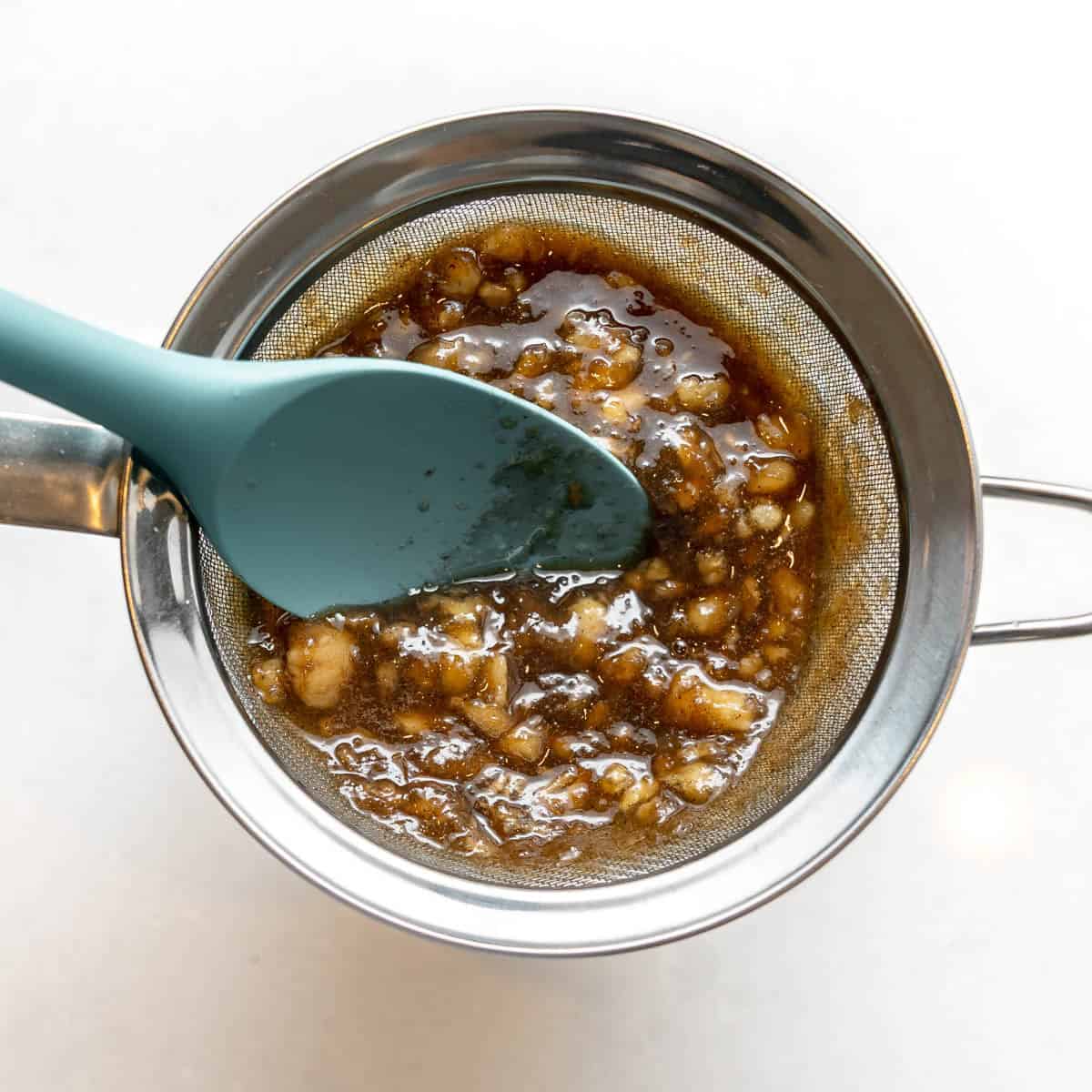 A metal strainer over a bowl with a brown, chunky liquid being pressed through it with a blue silicone spatula.