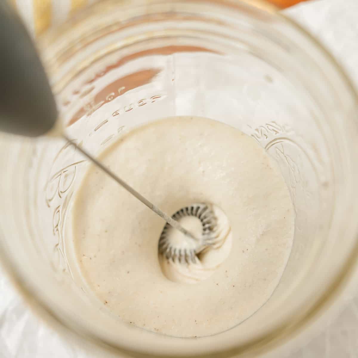 A handheld milk frother is mixing a creamy liquid in a glass jar, creating foam on the surface.