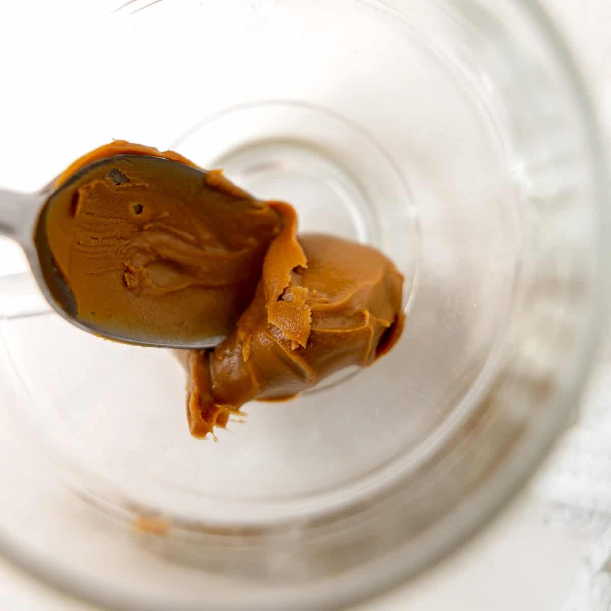 A spoon holding a dollop of brown peanut butter rests inside an empty, clear glass jar viewed from above.