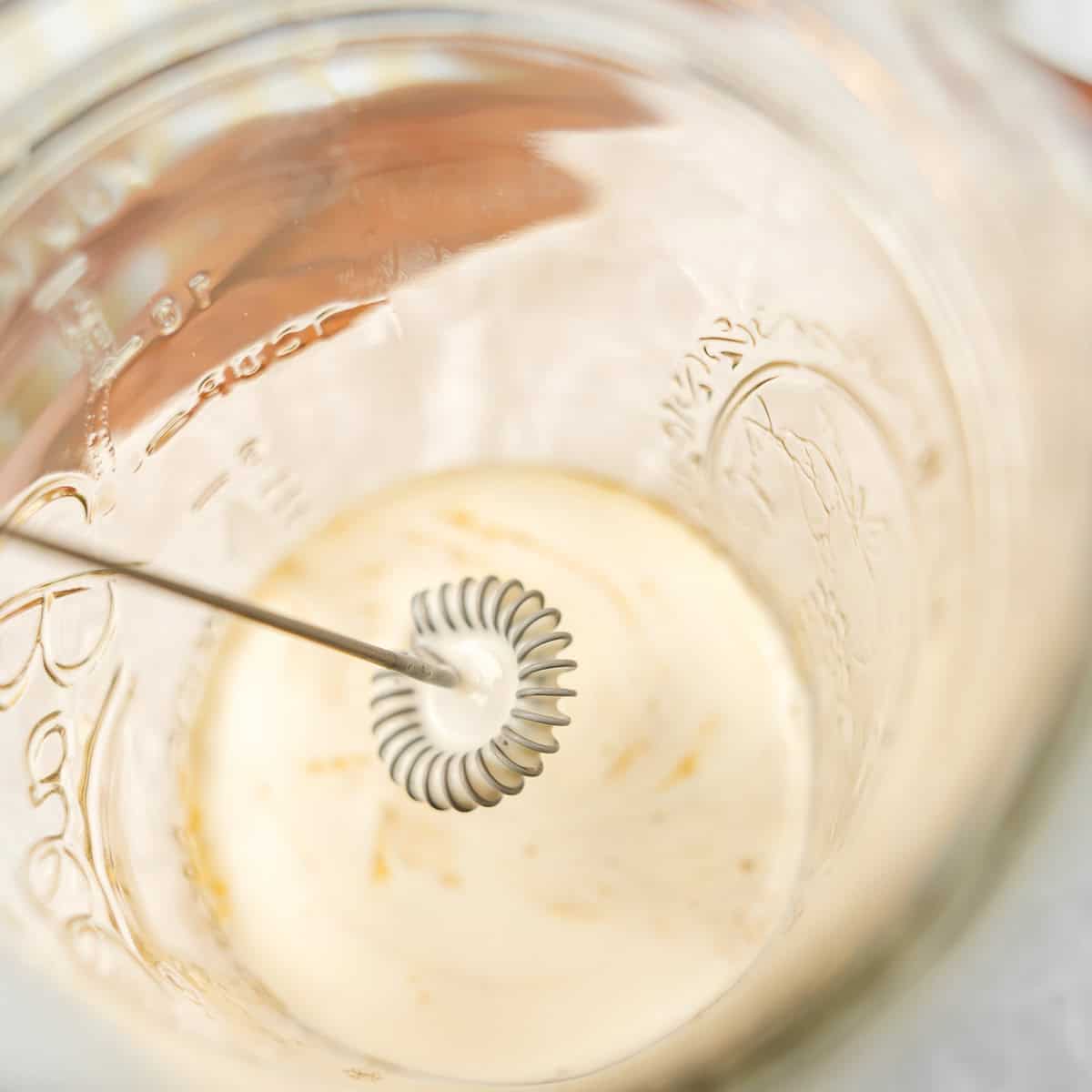 A close-up of a handheld milk frother whisking a small amount of liquid inside a glass jar.
