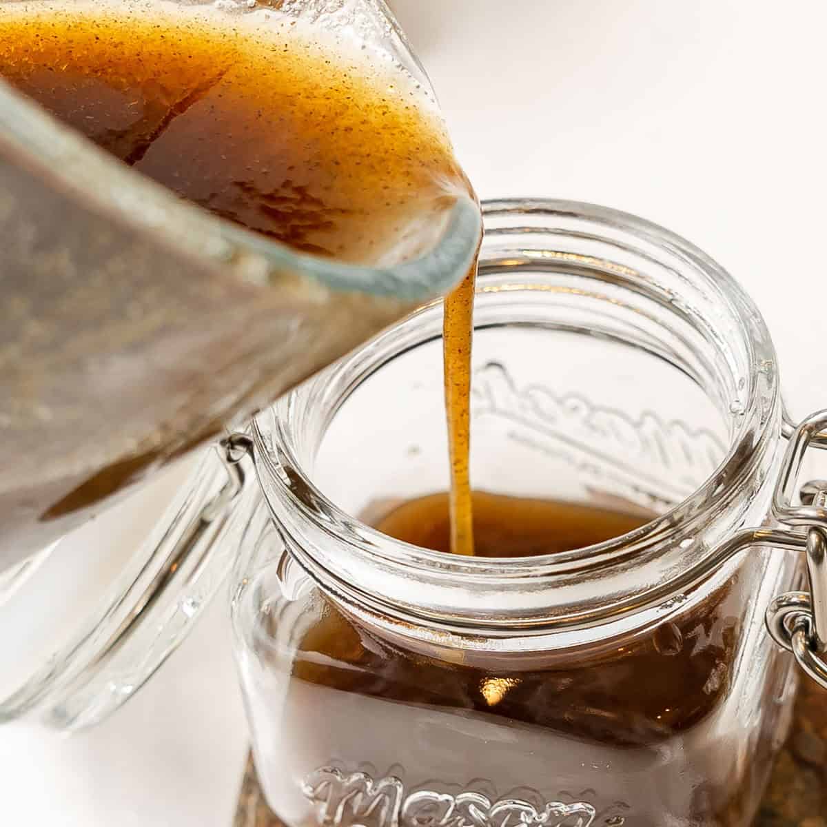 Brown liquid is being poured from a glass measuring cup into an open glass mason jar on a white surface.