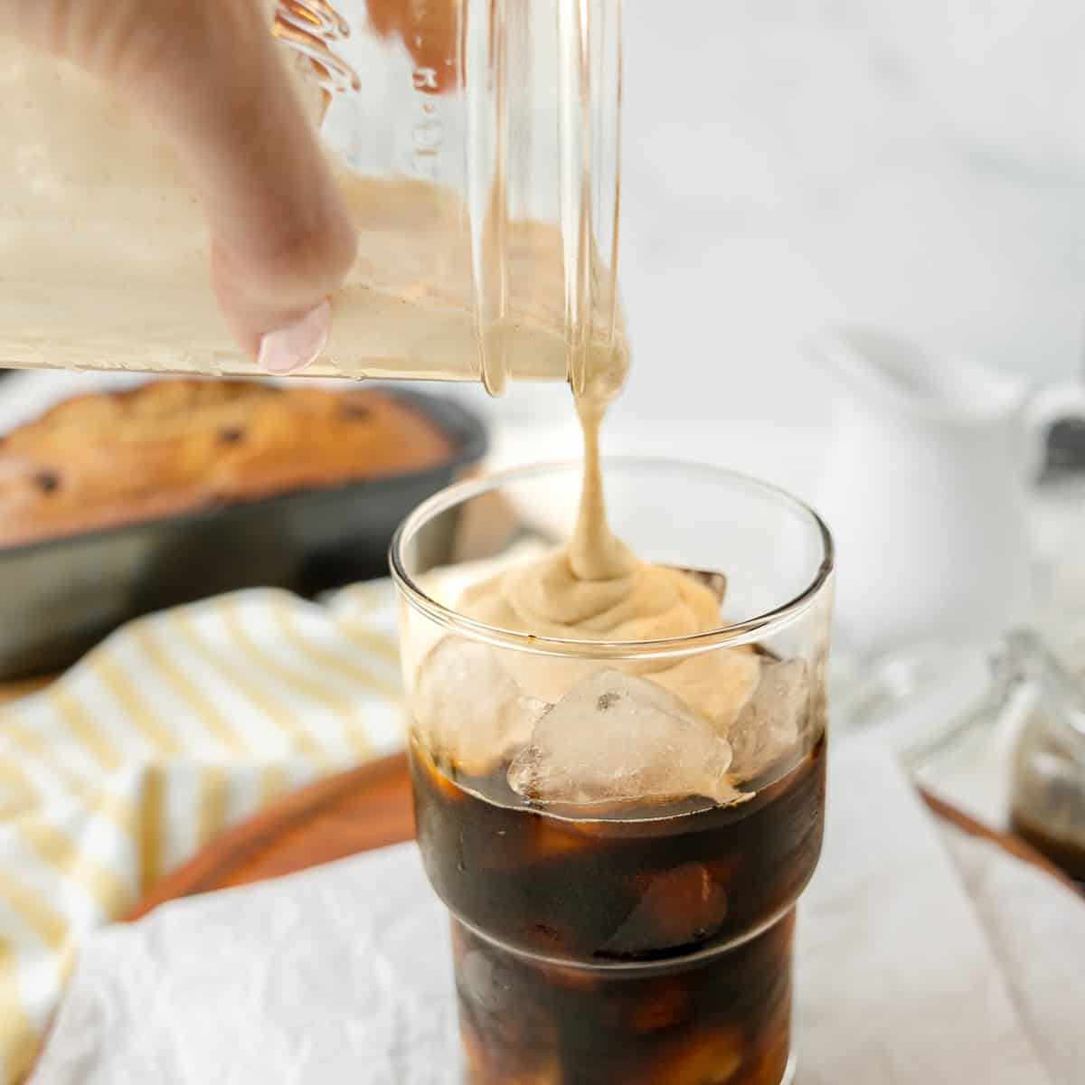 A hand pours creamy whipped coffee topping over a glass of iced coffee; a loaf of bread and striped towel are in the background.
